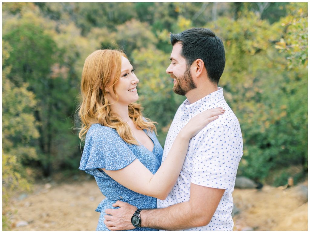 Engaged couple facing each other and smiling in front of a background of green trees
